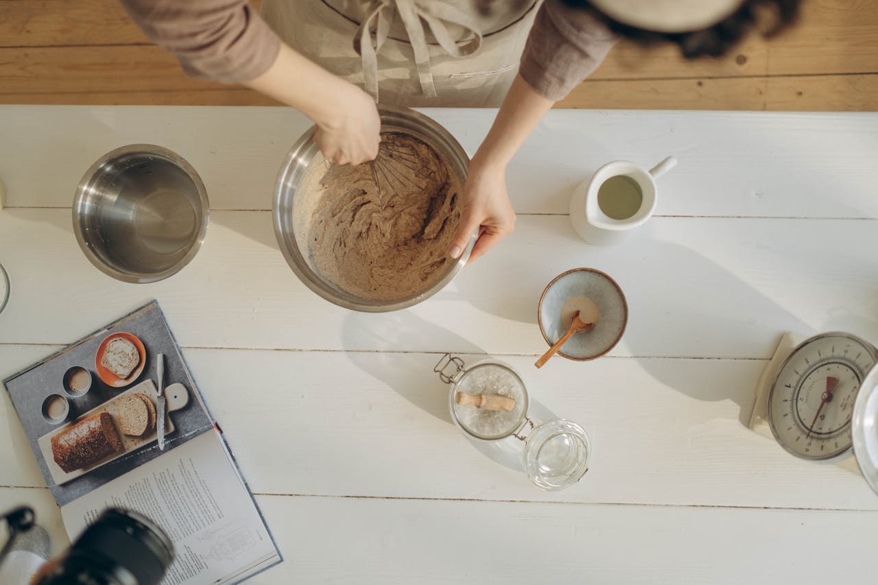 Home A close-up of hands mixing batter in a bowl among baking ingredients on a wooden table.